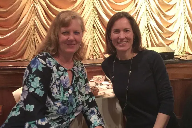 صورة لامرأتين تجلسان معًا في مكان مغلق، مع خلفية من الستائر، تعكس أجواء اجتماعية.