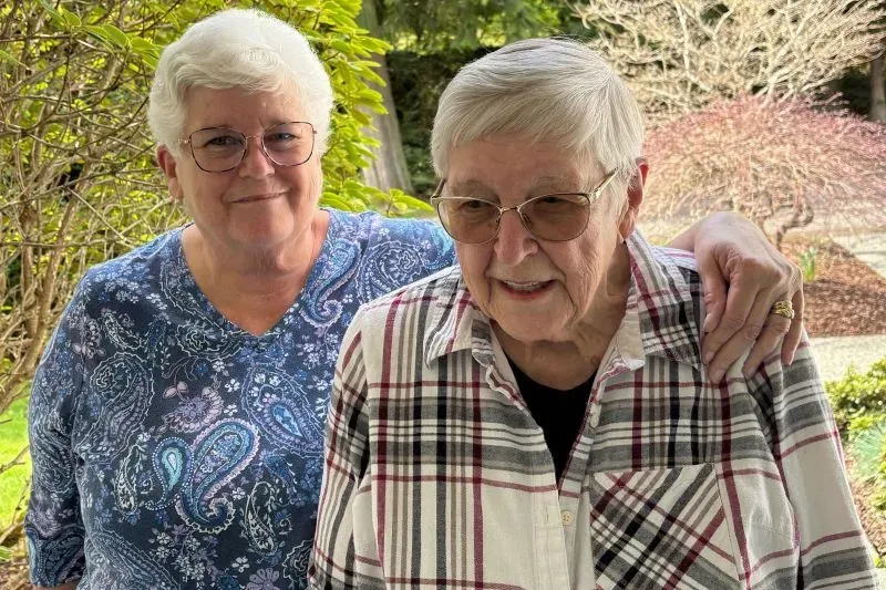 امرأتان مسنّتان مبتسمتان، واحدة ترتدي قميصًا مزخرفًا والأخرى بلوزة مخططة، يقفن معًا في حديقة، تعكسان قلق المستفيدين من خدمات الضمان الاجتماعي.