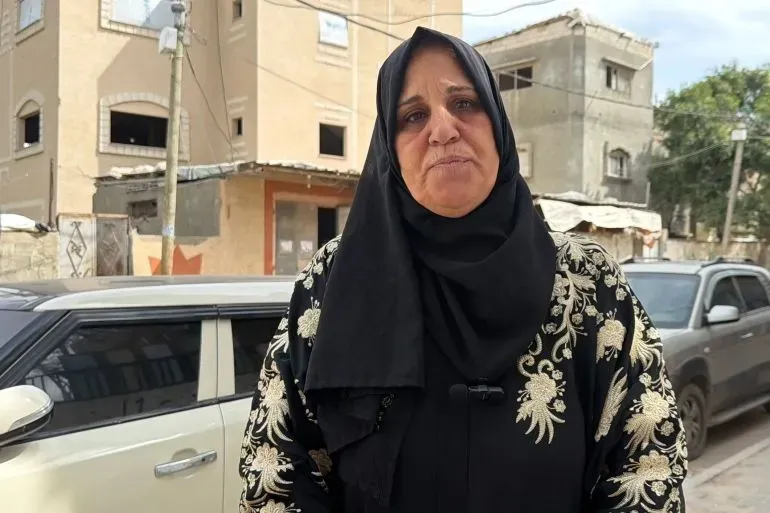 امرأة ترتدي عباءة سوداء وتظهر تعابير القلق، تقف في شارع دير البلح، خلفها مباني متضررة، تعكس آثار الحرب والانتخابات البلدية المقبلة.