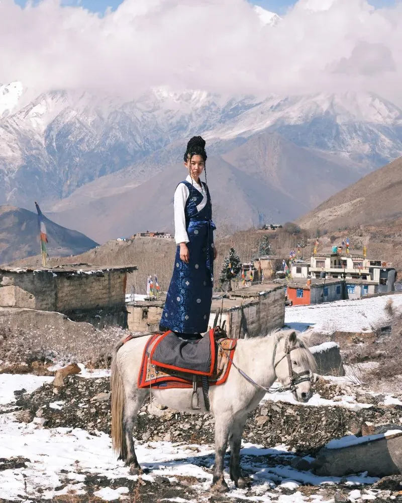 امرأة ترتدي زيًا تقليديًا تقف على ظهر حصان أبيض في منطقة جبلية مغطاة بالثلوج، مع منازل خلفها.