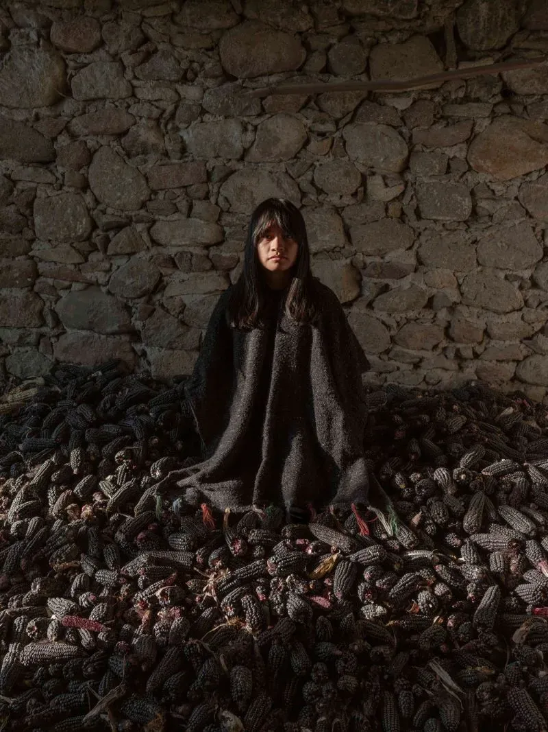 امرأة تجلس وسط كومة من حبوب الذرة السوداء في مكان ذو جدران حجرية، تعكس صورة من الحياة التقليدية والتراث الثقافي.