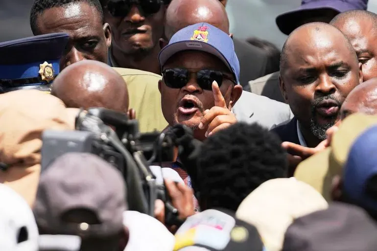 وزير الشرطة السابق سينزو متشونو يتحدث بحماس وسط حشد من الناس، مع وجود قوات أمنية في الخلفية، في سياق مزاعم فساد.