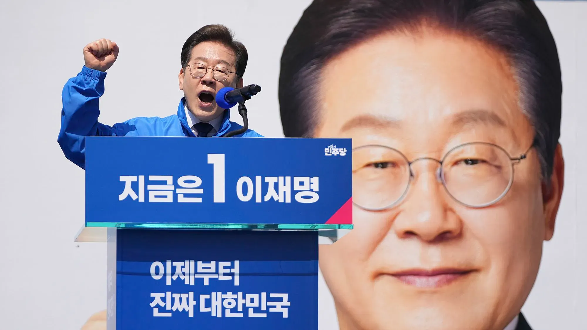 لي جاي ميونج، زعيم المعارضة في كوريا الجنوبية، يتحدث بحماس خلال حملته الانتخابية، مع صورة كبيرة له خلفه.