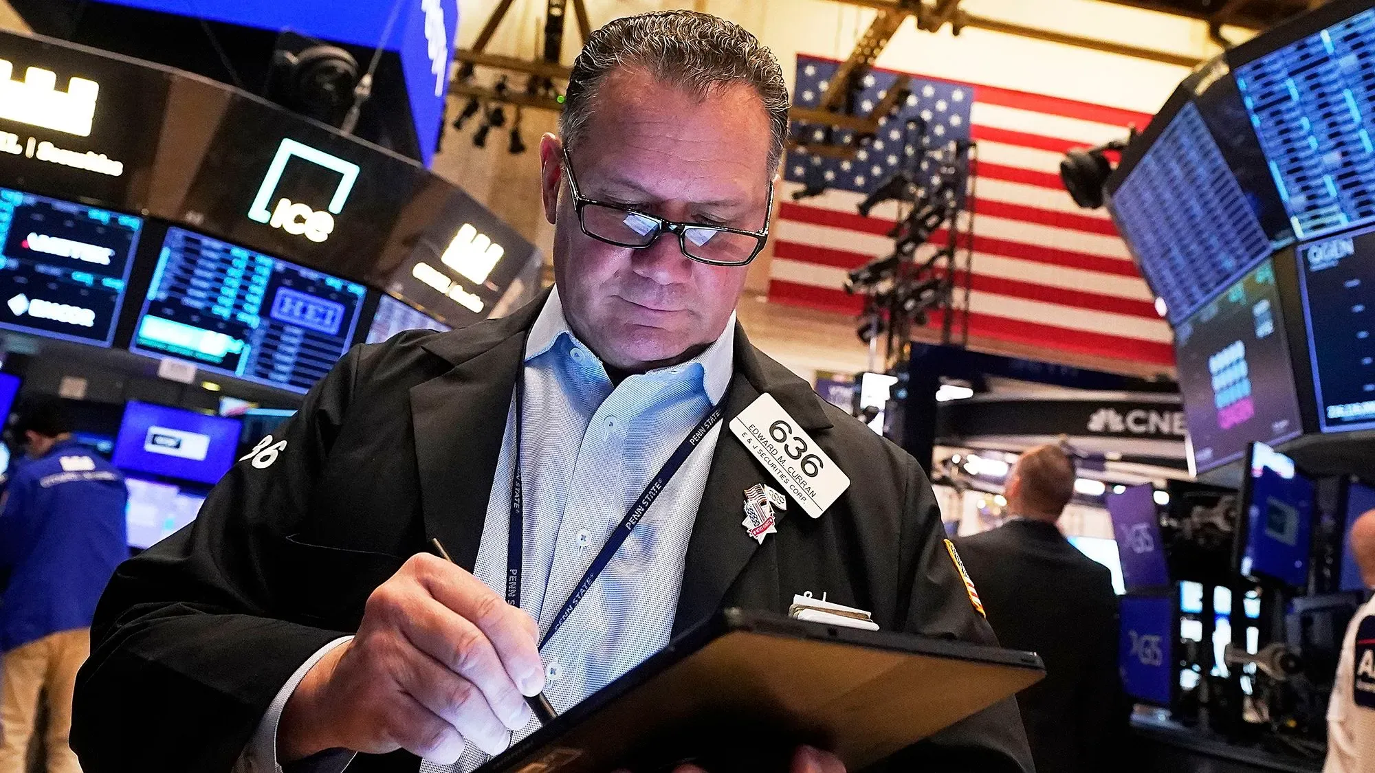 تاجر في بورصة نيويورك يتابع الأسهم ويكتب ملاحظات، مع ظهور شاشات التداول والخلفية الوطنية الأمريكية.