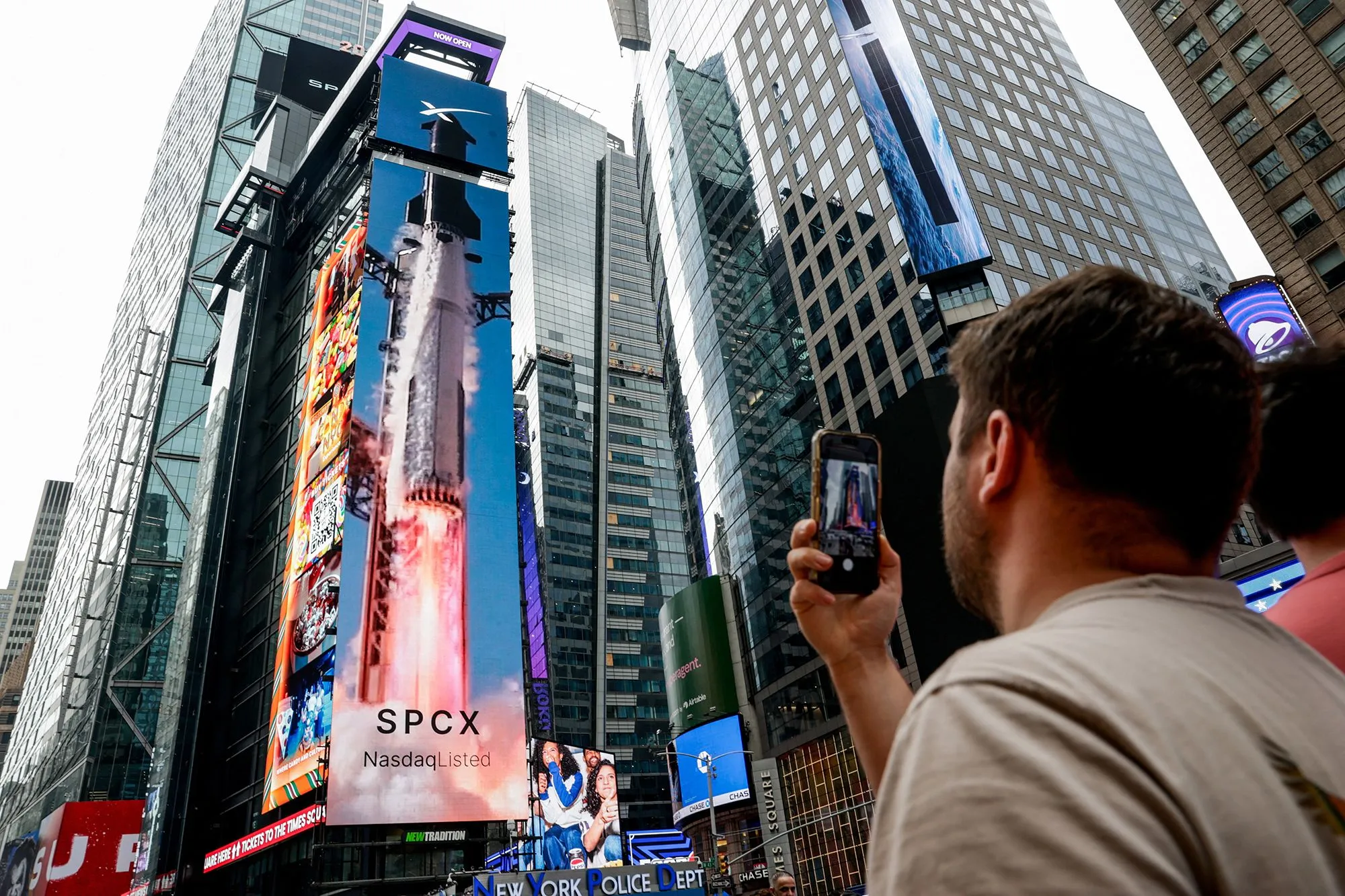 شخص يلتقط صورة لهاتفه لعرض SpaceX الضخم في تايمز سكوير، الذي يبرز تقييم الشركة المرتفع وأحدث صفقة استحواذ لها.