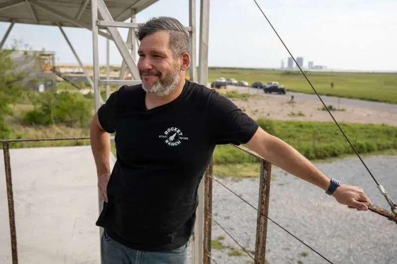 رجل يقف مبتسمًا في موقع بالقرب من سبيس إكس، مع منظر لحقول مفتوحة ومرافق الإطلاق في الخلفية، في منطقة ستاربايس بتكساس.