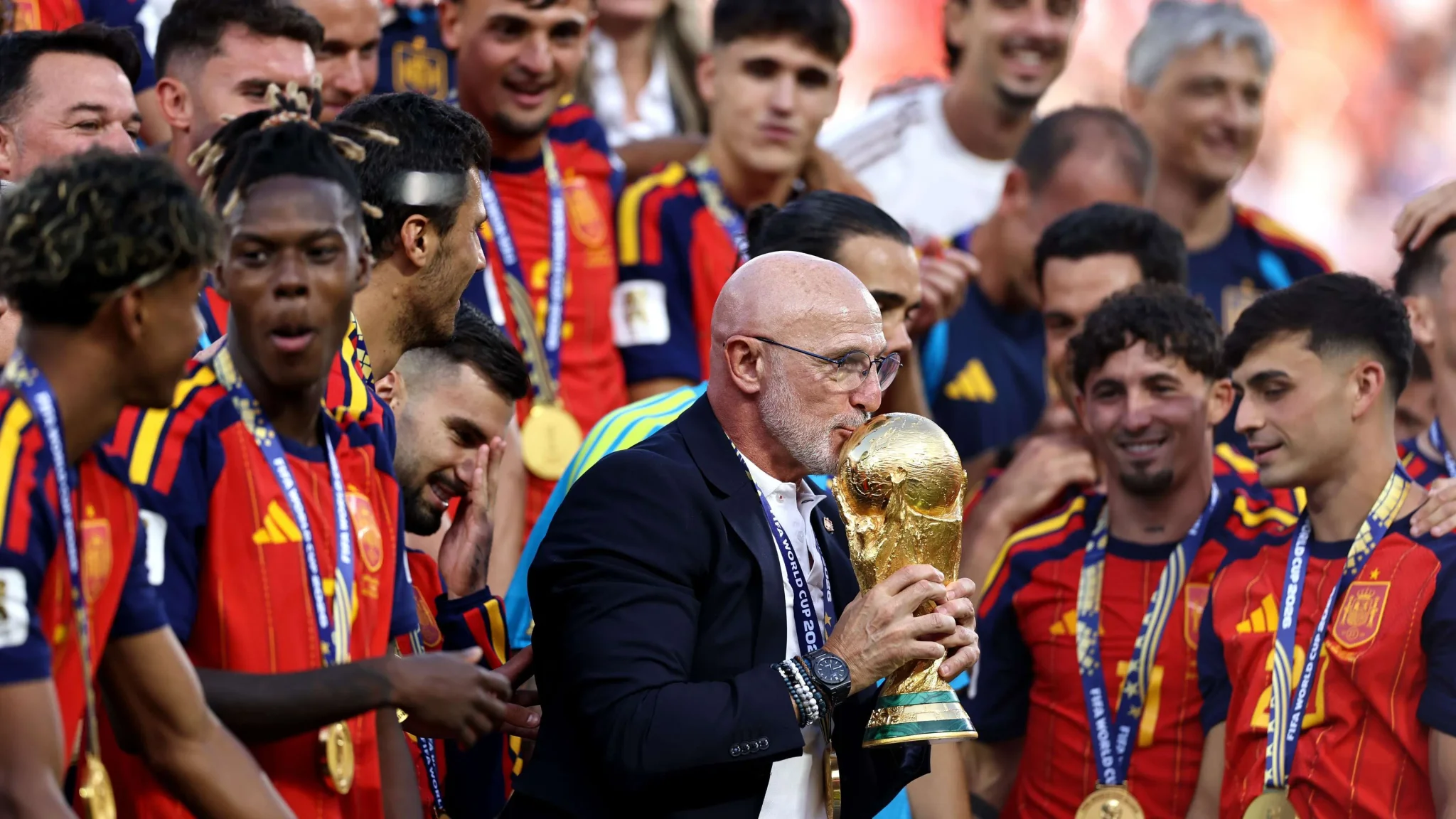 مدرب منتخب إسبانيا يقبل كأس العالم وسط لاعبين مبتسمين يرتدون زي الفريق الأحمر في احتفال الفوز بكأس العالم 2026.