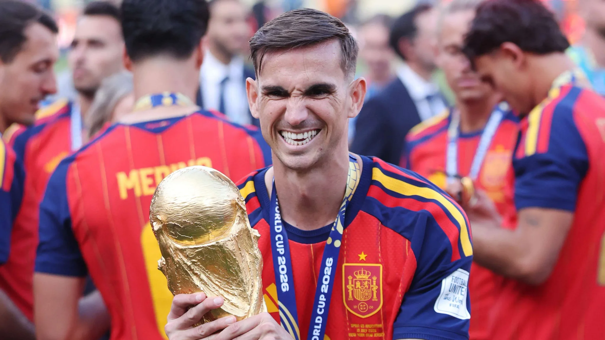لاعب منتخب إسبانيا يحمل كأس العالم 2026 مبتسمًا بفخر وسط زملائه، مع أجواء احتفالية بعد الفوز بالبطولة.