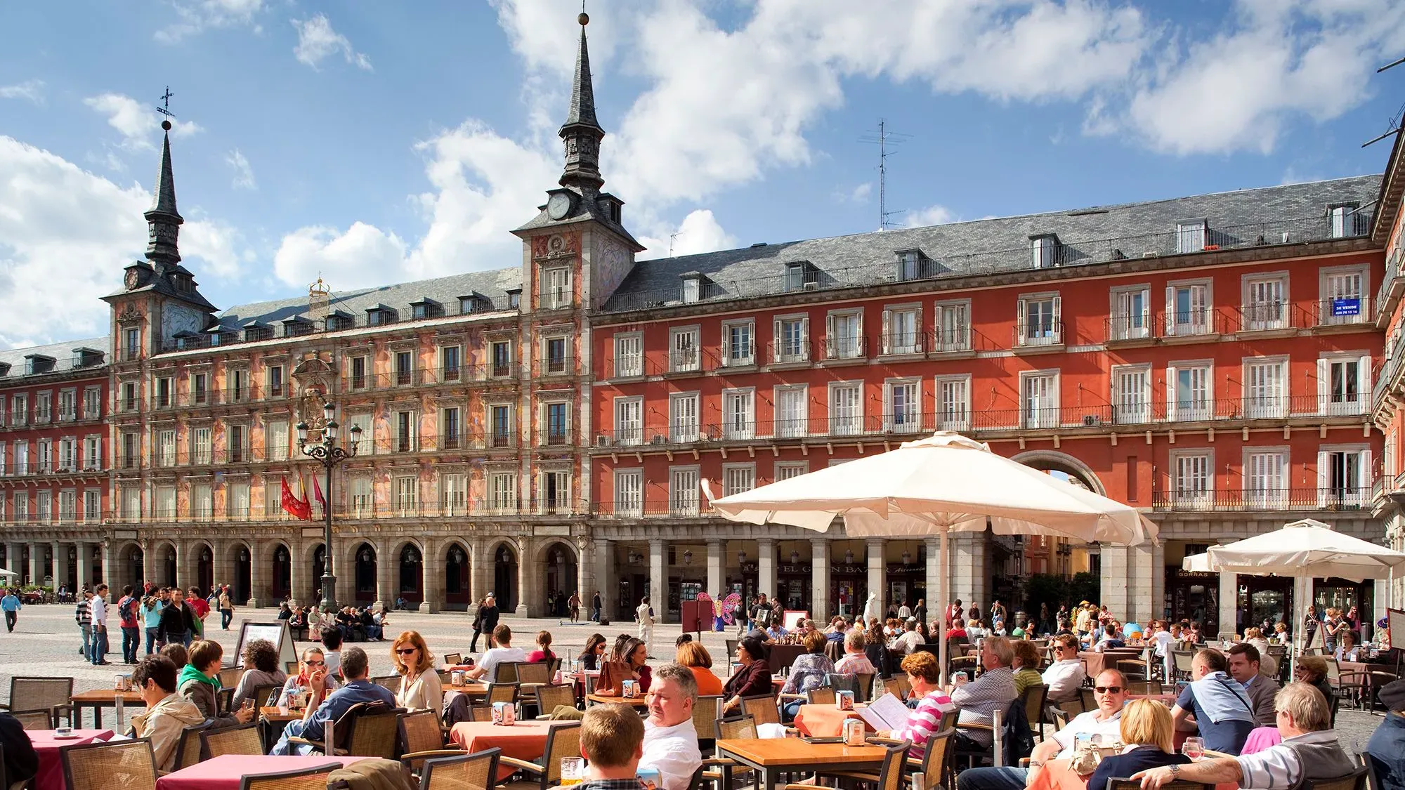 مجموعة من الزوار يستمتعون بالغداء في مطعم خارجي في ساحة بلازا مايور بمدريد، تظهر العمارة الأنيقة والسماء الزرقاء.