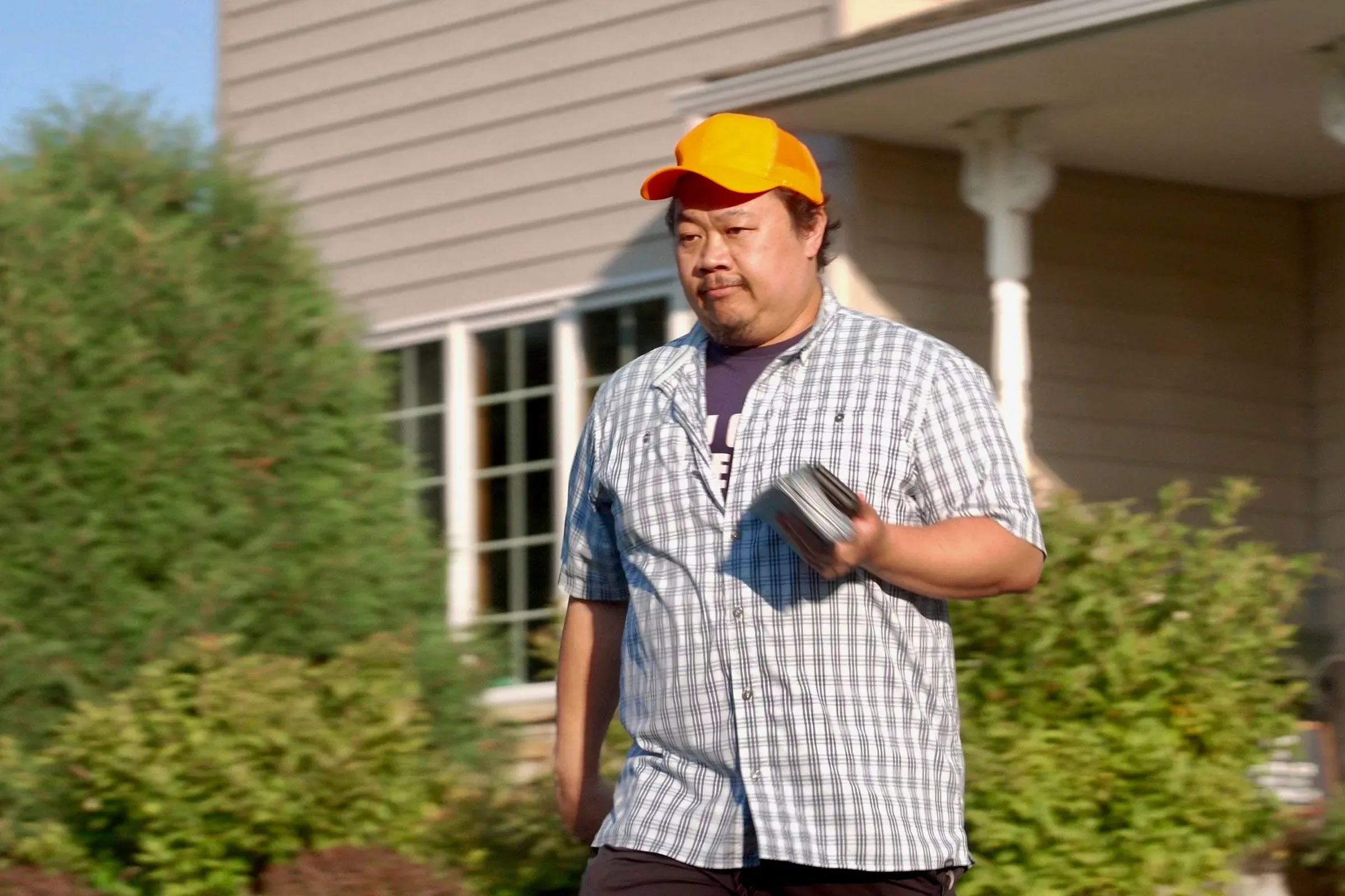 رجل يرتدي قبعة برتقالية وقميصًا مخططًا يسير في حي سكني، يحمل هاتفًا محمولًا، وسط أجواء انتخابية في مينيسوتا.