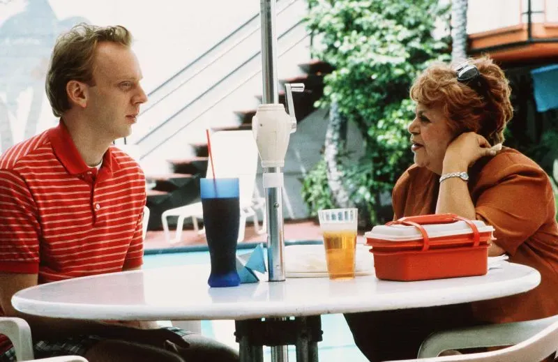 مشهد من فيلم يجمع شخصيتين تتناولان المشروبات على طاولة بجوار المسبح، مع خلفية خضراء وأجواء غير رسمية.