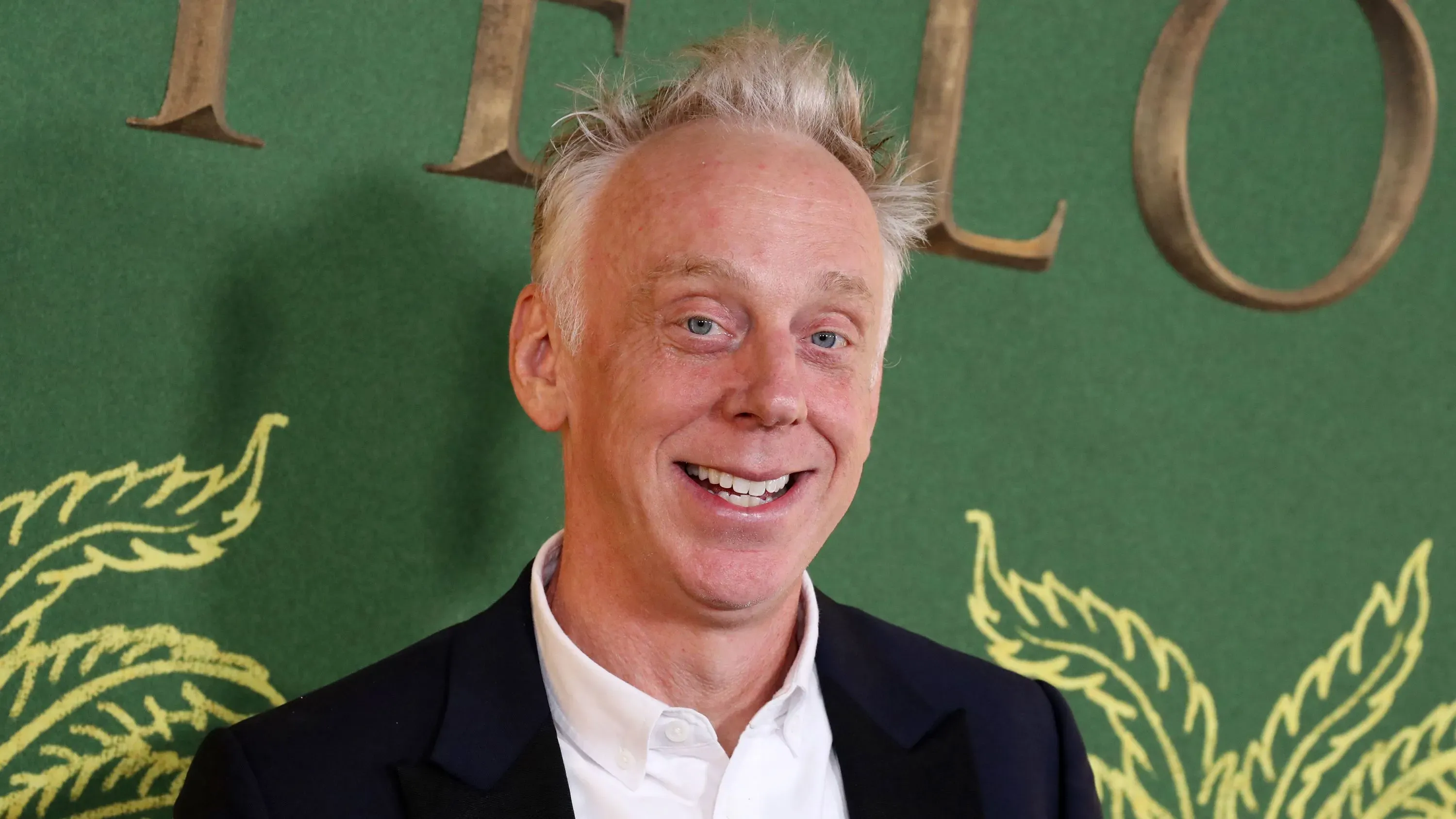 مايك وايت مبتسم في حدث رسمي، يرتدي بدلة سوداء مع قميص أبيض، خلفه خلفية خضراء تحمل تصميم نباتي، يعكس نجاحه في هوليوود.