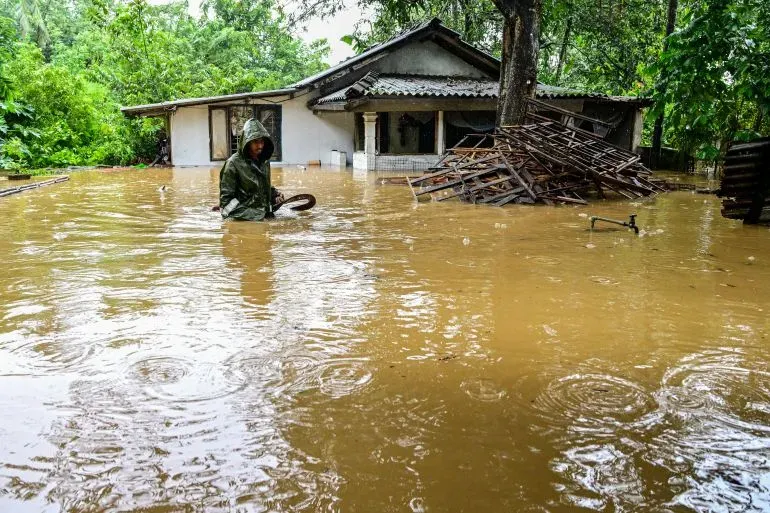 غمر الفيضانات منطقة سكنية في سريلانكا، حيث يظهر شخص يرتدي معطفاً واقياً وسط المياه، مع منزل متضرر في الخلفية.