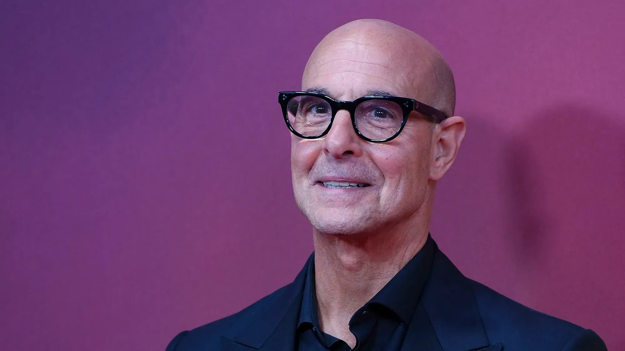 ستانلي توتشي مبتسم يرتدي نظارات، يتحدث عن تجربته مع علاج السرطان وتأثيره على صحته، في خلفية ملونة.