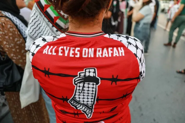قميص أحمر يحمل عبارة "كل الأنظار على رفح" مع رسومات تعكس الوضع في غزة، وسط تجمع شعبي للتضامن مع الفلسطينيين.