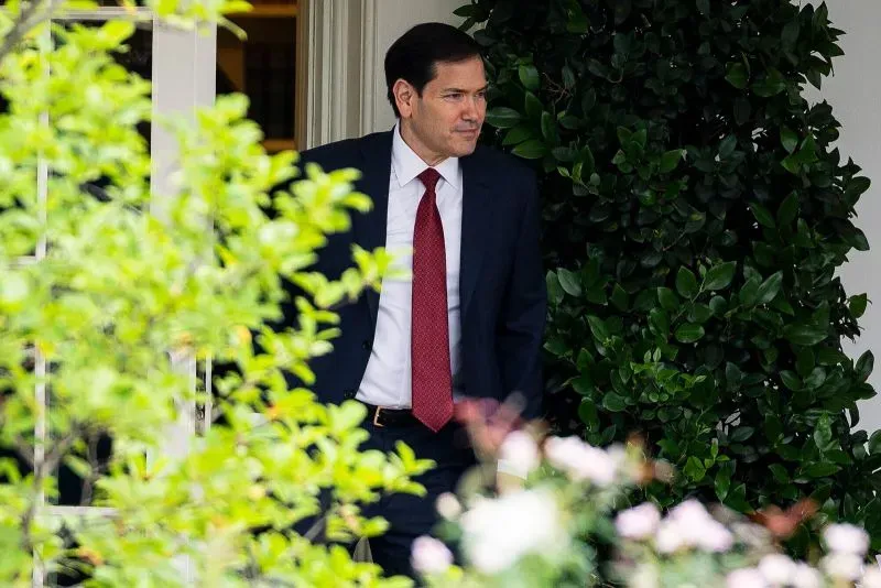 ماركو روبيو يظهر وهو يغادر مبنى حكومي، مع خلفية من النباتات الخضراء، في سياق مناقشات حول تخفيضات وزارة الخارجية.