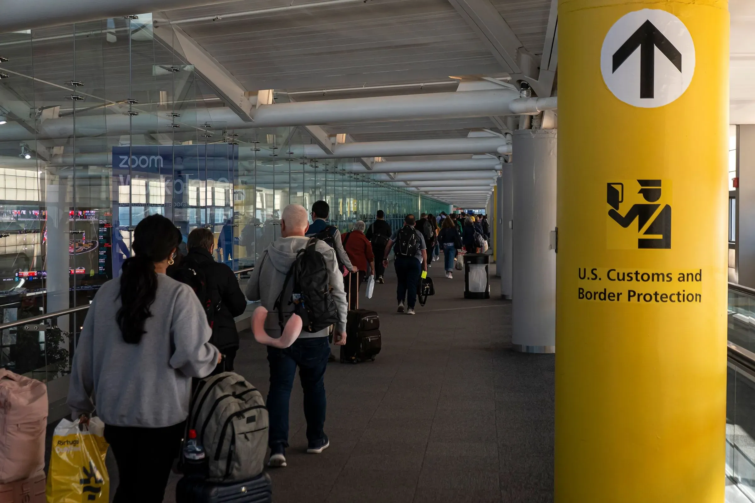 طابور من المسافرين في مطار أمريكي، متجهين نحو نقطة تفتيش الجمارك وحماية الحدود، مع لافتة تشير إلى الاتجاه.