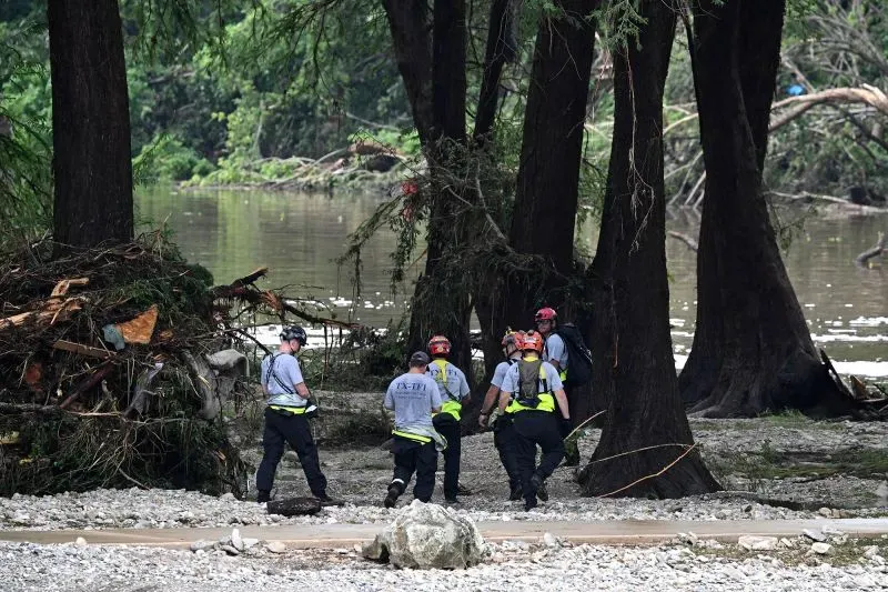 فرق الإنقاذ تعمل في موقع مخيم ميستيك بعد الفيضانات، مع وجود حطام وأشجار قرب نهر غوادالوبي، مما يعكس الأضرار الكبيرة.