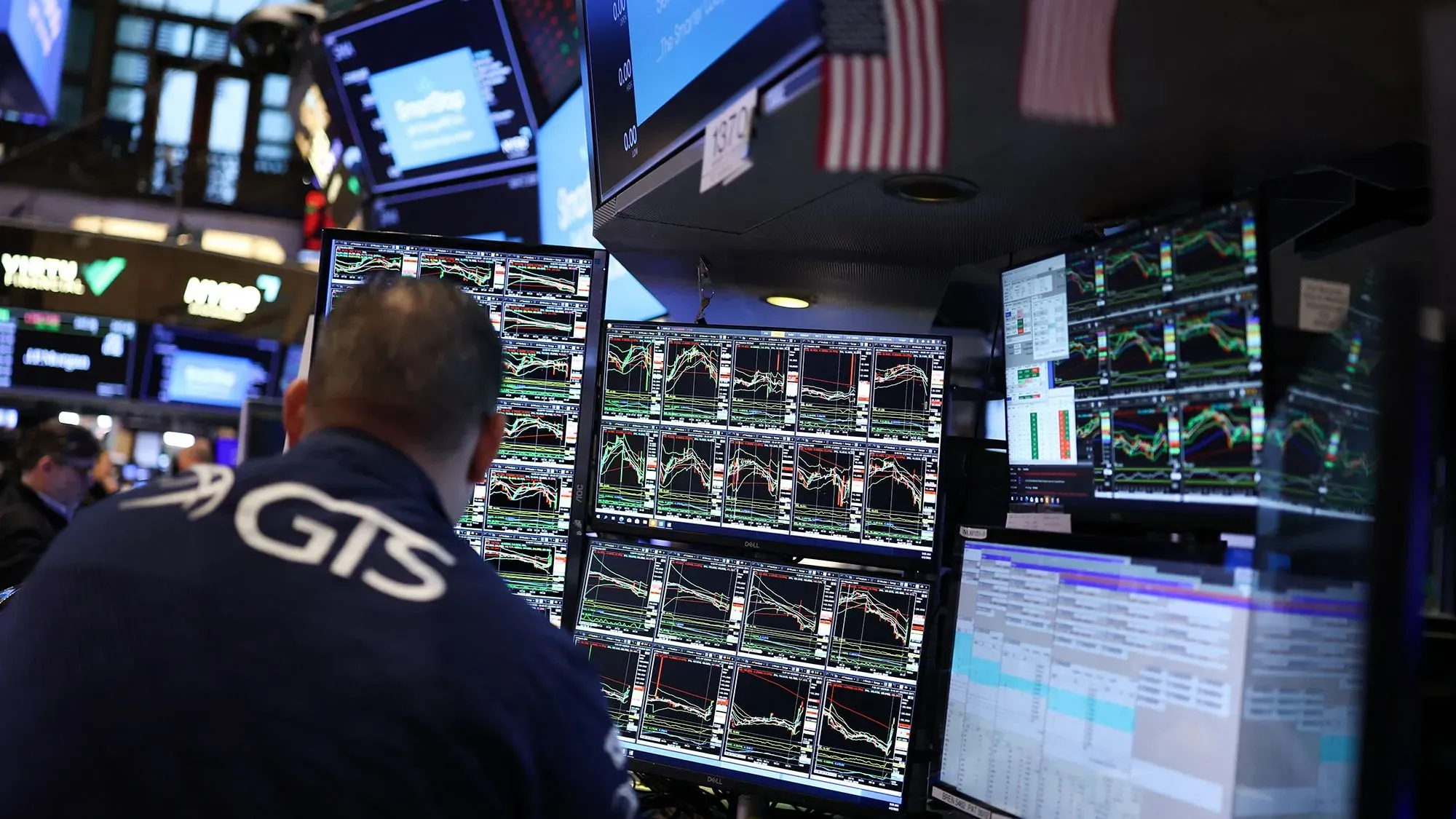 تظهر الصورة تاجرًا في سوق الأسهم محاطًا بشاشات تعرض بيانات السوق، مما يعكس تأثير الرسوم الجمركية على الاقتصاد.