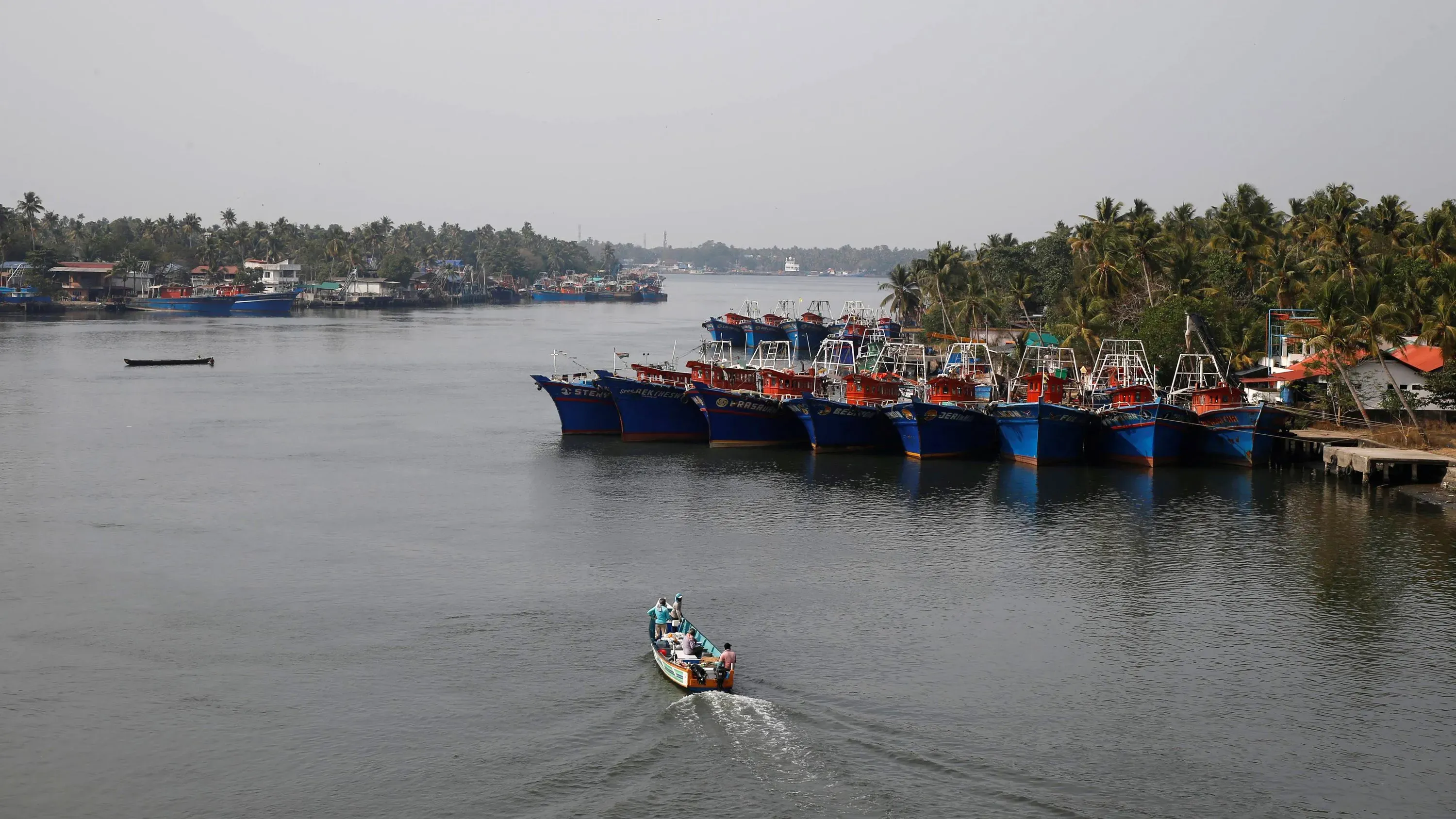 قارب صغير يبحر في قناة مائية محاطة بسفن صيد ملونة وأشجار جوز الهند في ولاية كيرالا، مشهد يعكس جمال السياحة والثقافة المحلية.