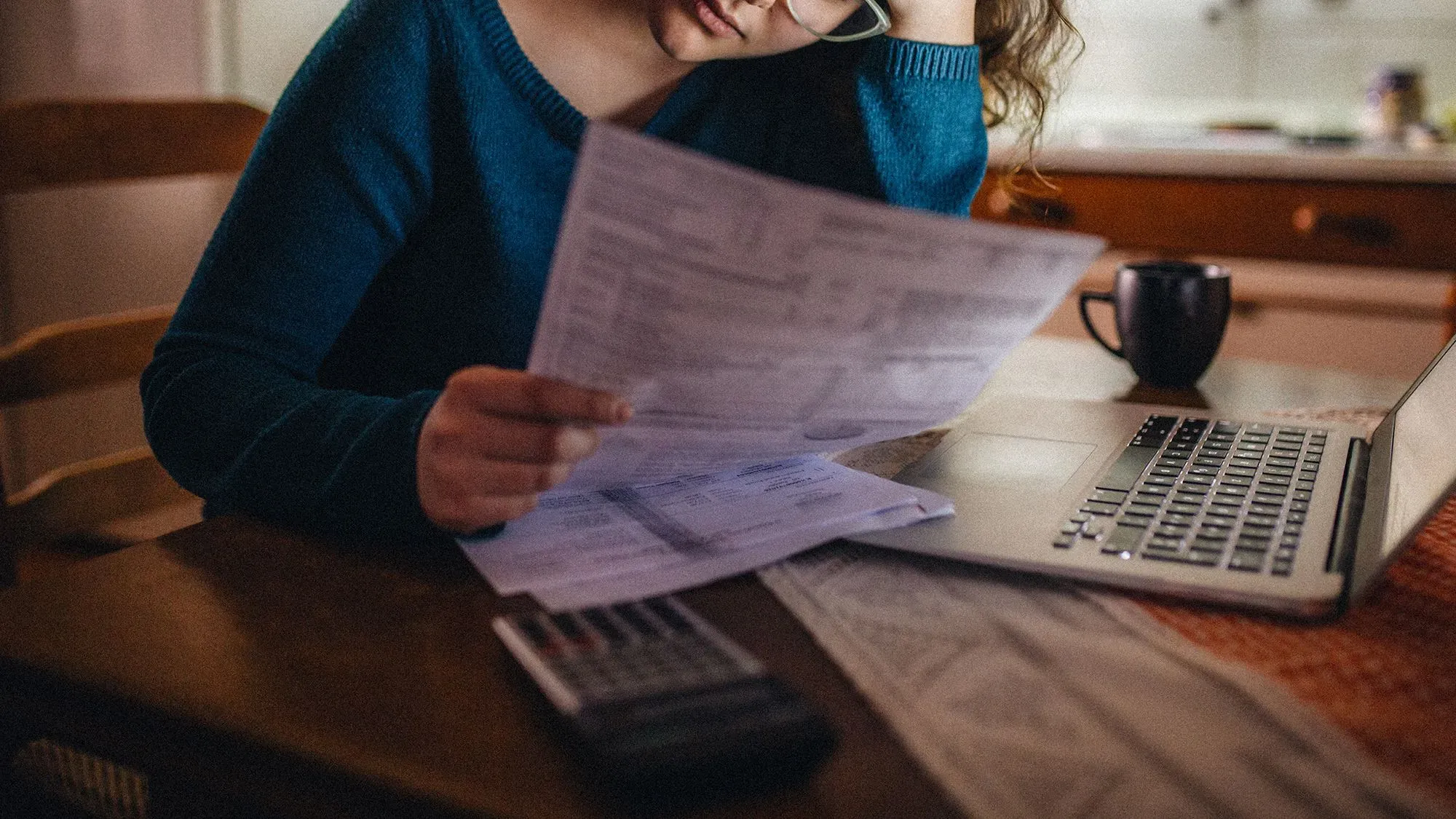 امرأة ترتدي سترة زرقاء تجلس أمام مكتب، تمسك بأوراق مالية وتبدو قلقة، مع حاسوب محمول وآلة حاسبة أمامها.