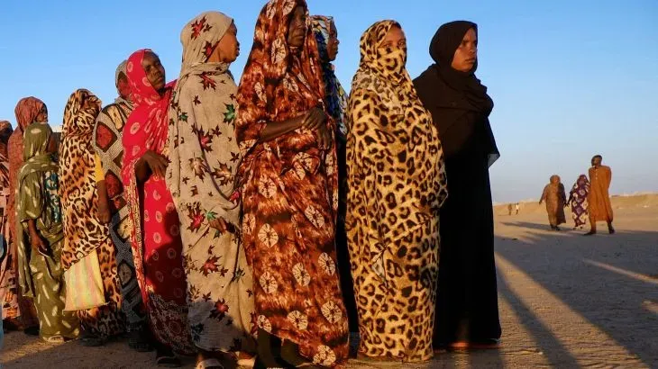 نساء يرتدين أزياء تقليدية، يقفن في صف طويل تحت ضوء الشمس، تعبيرات وجوههن تعكس المعاناة خلال فرارهن من الفاشر في دارفور.