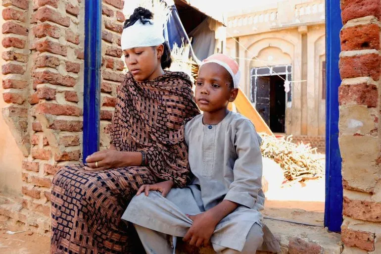 امرأة وطفل يجلسان معًا في منطقة متضررة من النزاع في السودان، يظهر على رأس المرأة ضمادة، مما يعكس آثار الحرب المستمرة.