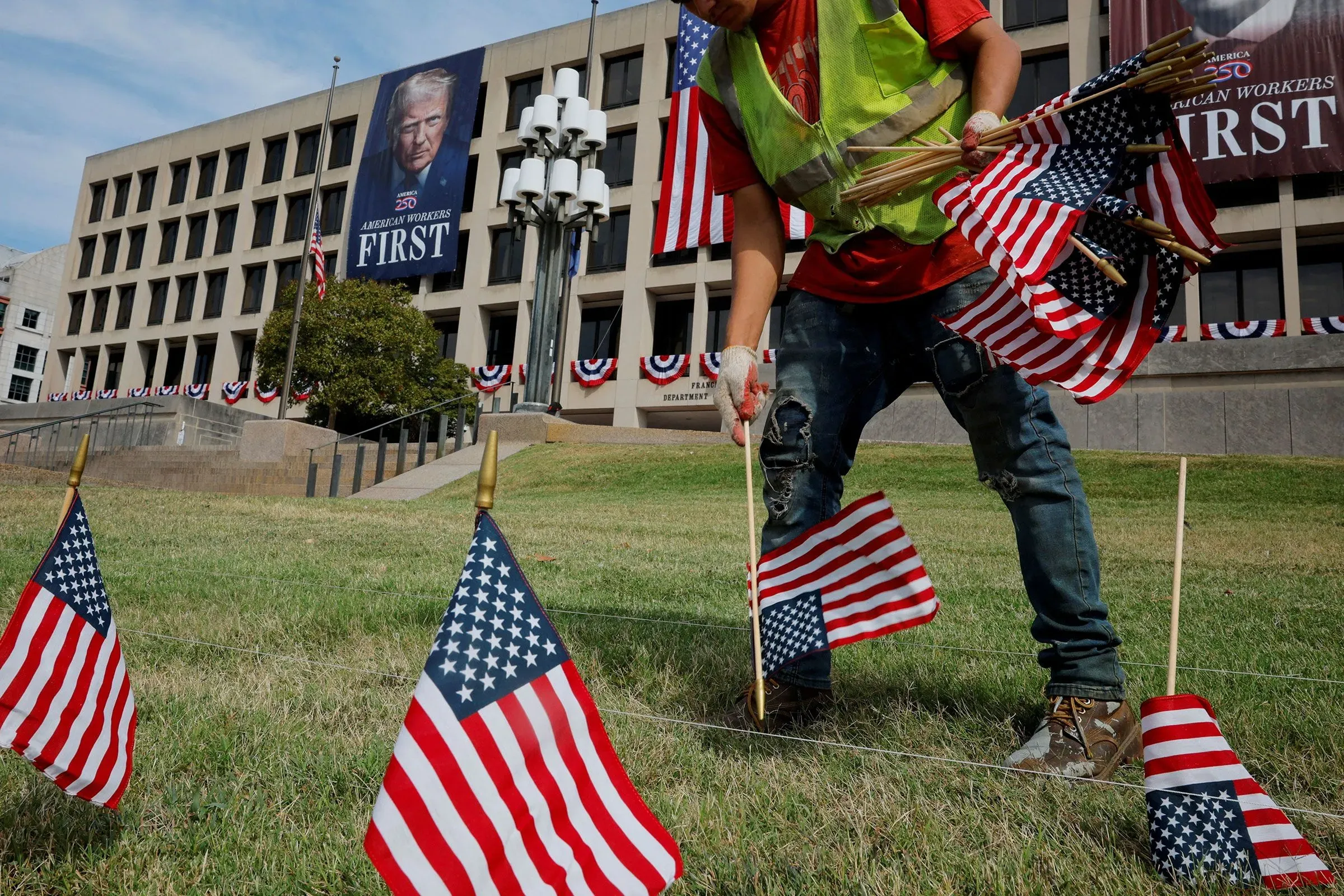 عامل يزرع أعلامًا أمريكية صغيرة أمام مبنى حكومي مزين بشعار "العمال الأمريكيون أولاً" بمناسبة عيد العمال.
