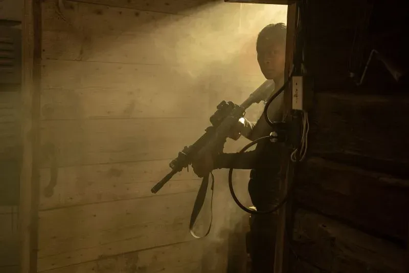 شخص يحمل سلاحًا في مكان مظلم، محاطًا بالدخان، مما يخلق جوًا من التوتر والإثارة، يتناسب مع أفلام الحركة والإثارة.