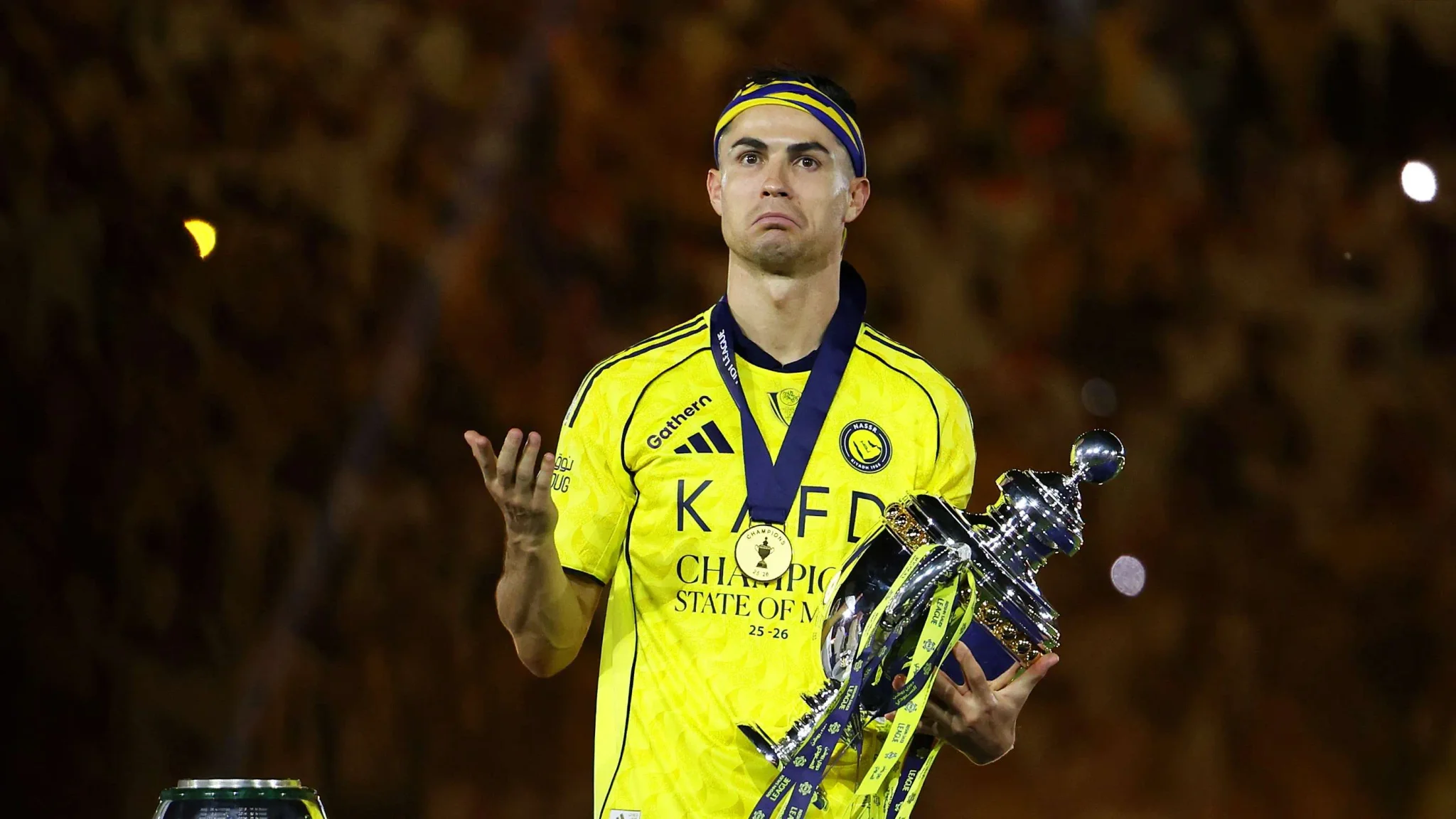 لاعب كرة قدم يرتدي زي نادي النصر السعودي ممسكًا بكأس البطولة مع تعبير وجه يعكس التحدي، في سياق موسم انتقالات دوري Roshn السعودي 2026.