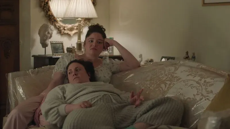 امرأتان تجلسان على أريكة في غرفة معيشة، تعبران عن مشاعر مختلفة أثناء مشاهدة التلفاز، تعكس لحظة من الاسترخاء والراحة.