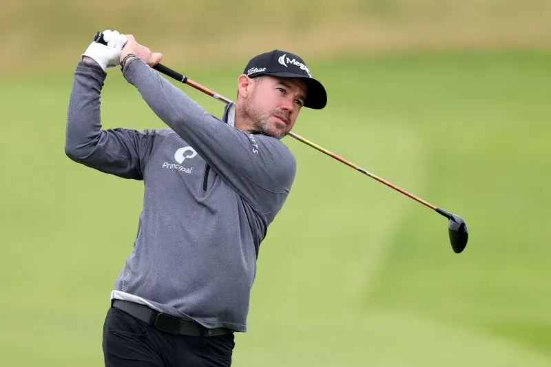 لاعب الجولف براين هارمان أثناء تسديد ضربة في ملعب جولف، يرتدي ملابس رياضية، مع خلفية طبيعية تُظهر المسطحات الخضراء.