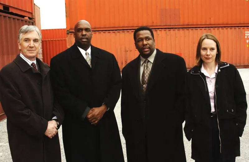 مجموعة من أربعة شخصيات رئيسية من مسلسل "ذا واير"، يقفون أمام حاويات شحن برتقالية، يعكسون أجواء درامية من العمل الشرطي.