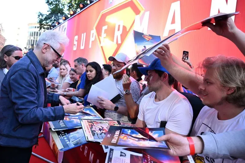 جيمس غان يوقع تذكارات لمشجعين خلال حدث ترويجي لفيلم "سوبرمان" الجديد، مع خلفية تحمل شعار الفيلم.
