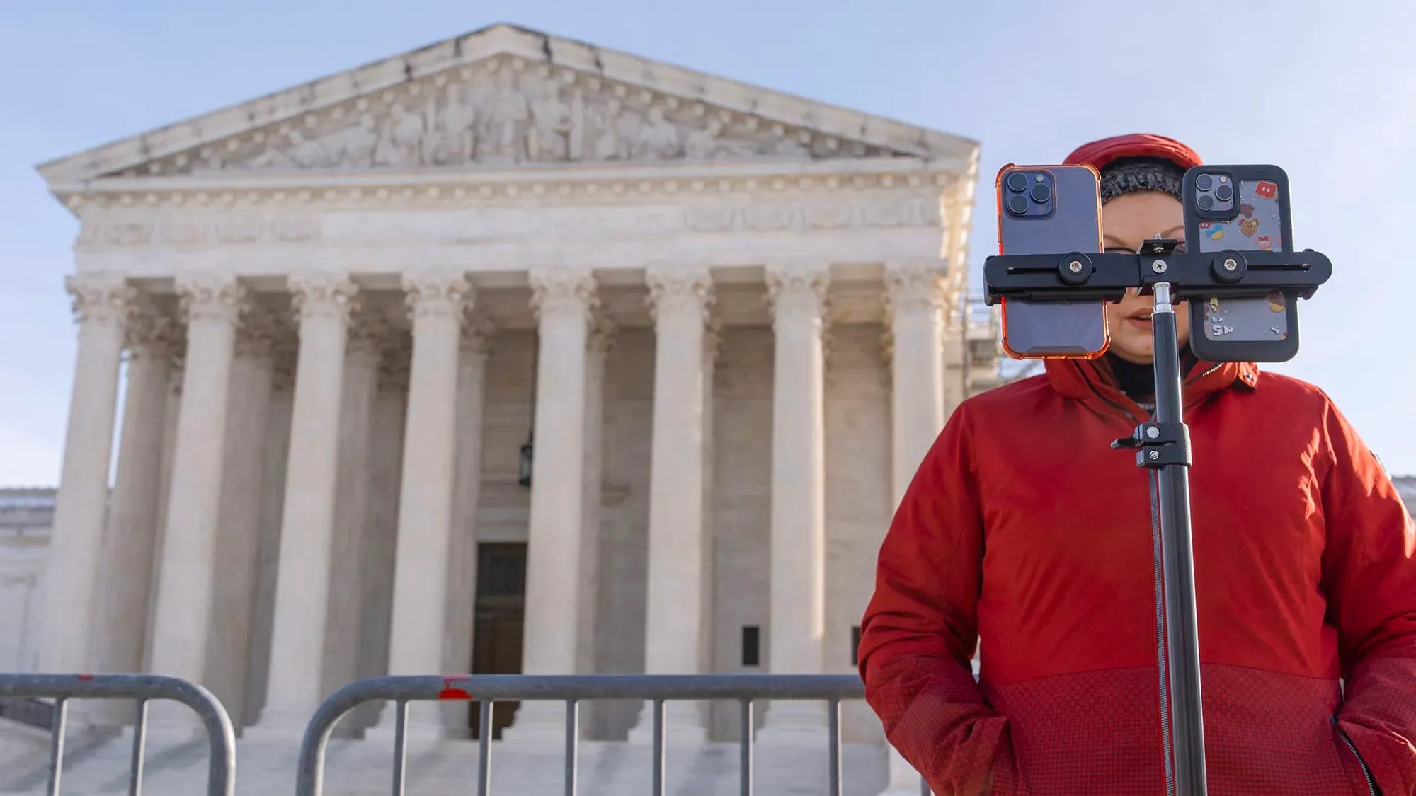 شخص يرتدي معطفًا أحمر ويستخدم حامل هواتف لتصوير نفسه أمام مبنى المحكمة العليا، وسط تحذيرات بشأن حظر TikTok.