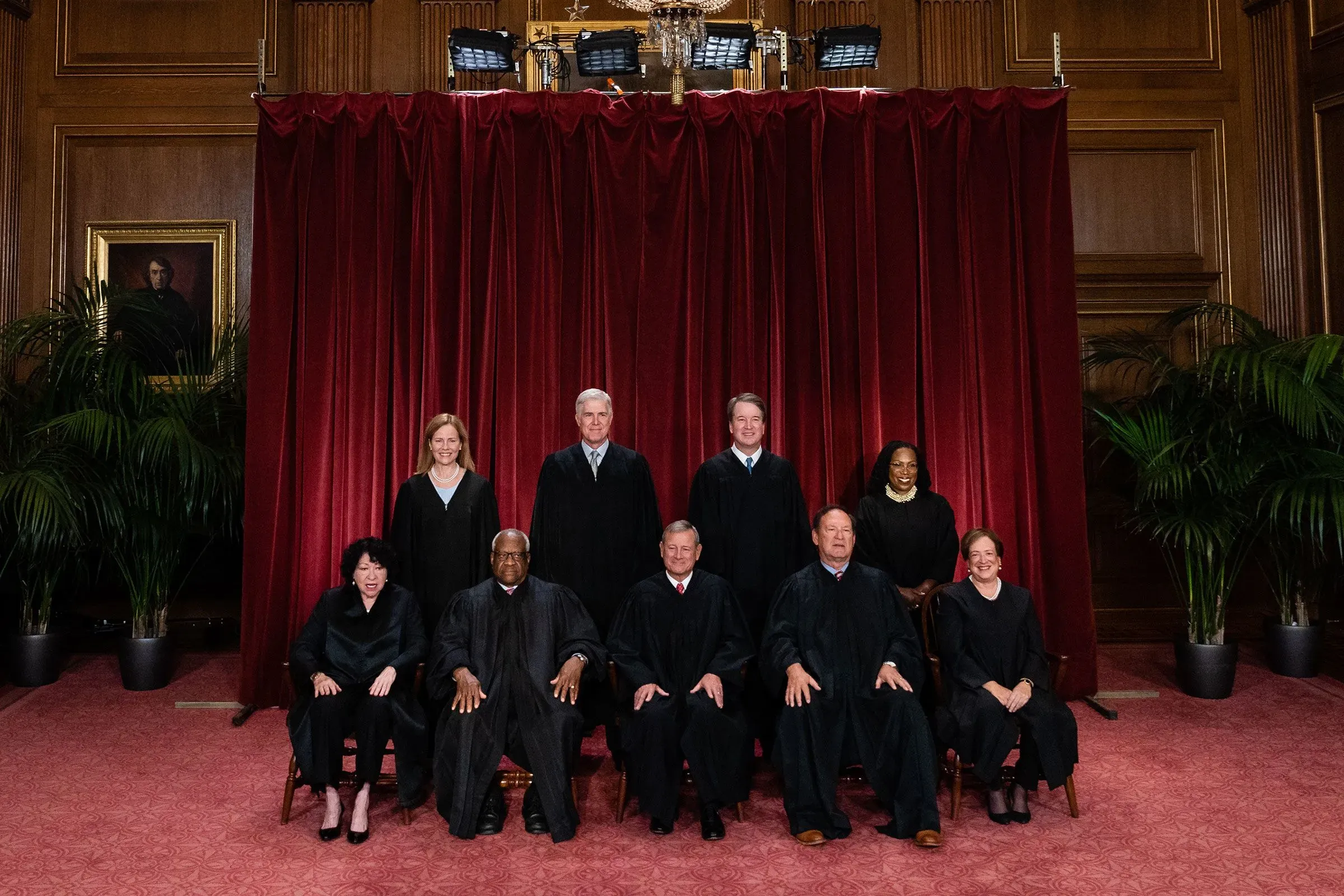 صورة جماعية لقضاة المحكمة العليا الأمريكية، مع خلفية حمراء، تظهر تسعة قضاة بملابس رسمية، تعكس التوترات السياسية الحالية في المحكمة.