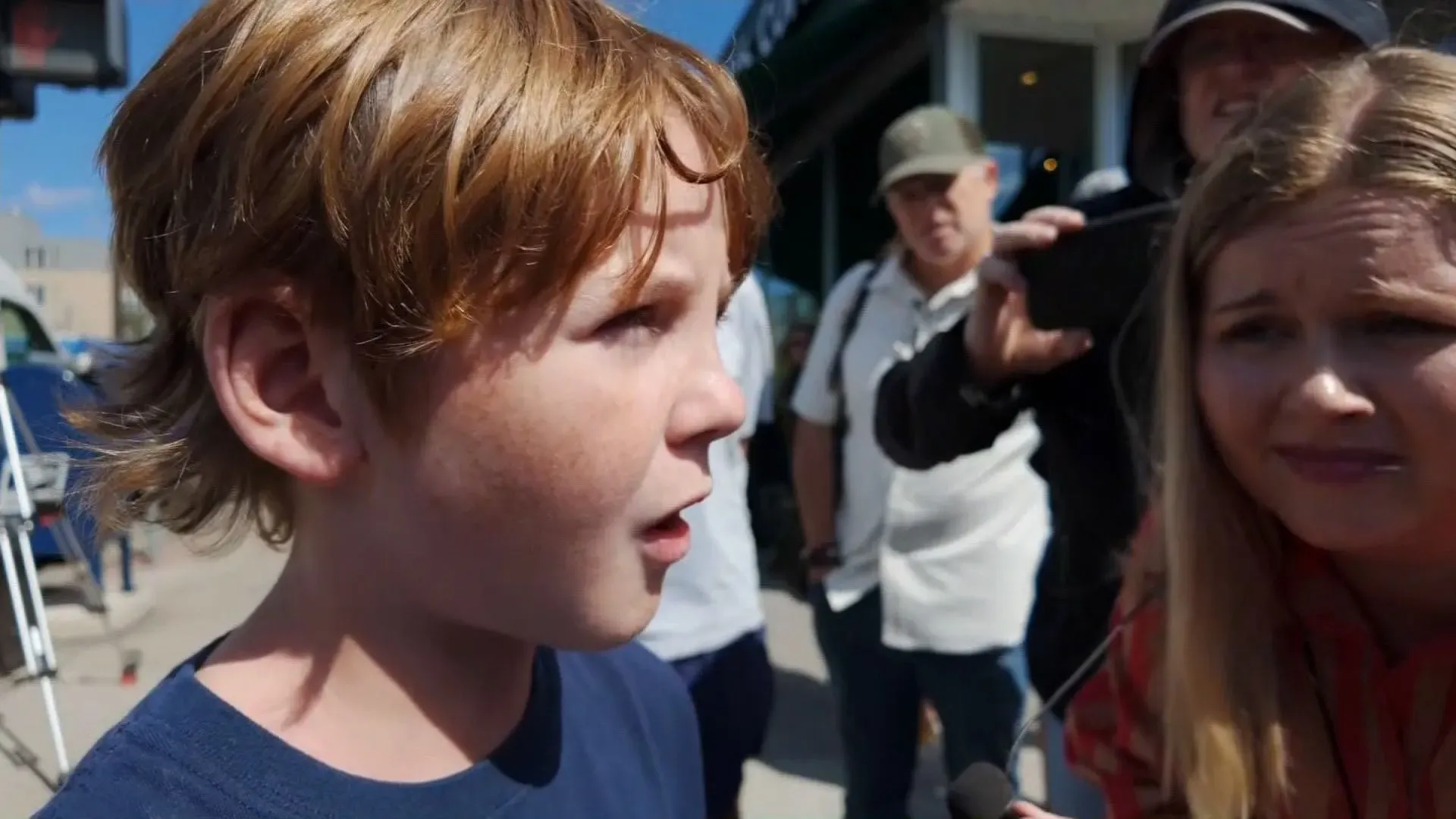 طفل يتحدث بقلق بعد إطلاق النار في مدرسة كاثوليكية، مع وجود أشخاص آخرين في الخلفية، يعكس تأثير الحادث على المجتمع.