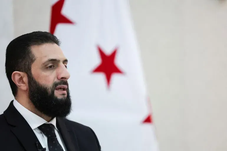 الرجل يتحدث بجدية أمام خلفية تحمل نجوم حمراء، في سياق مناقشات حول الأوضاع في سوريا والتهديدات الإسرائيلية.