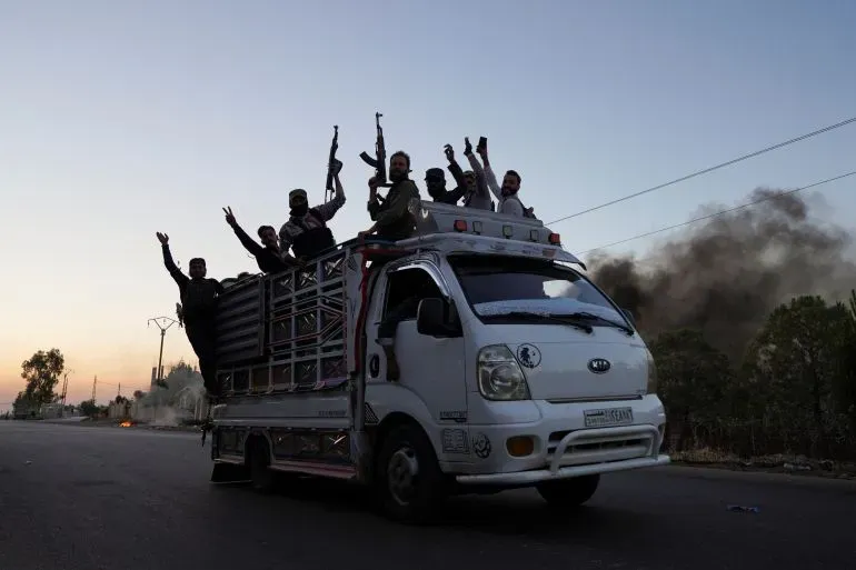 مقاتلون يحملون أسلحة يركبون شاحنة في محافظة السويداء السورية، مع تصاعد الدخان في الخلفية، وسط توترات أمنية متزايدة.