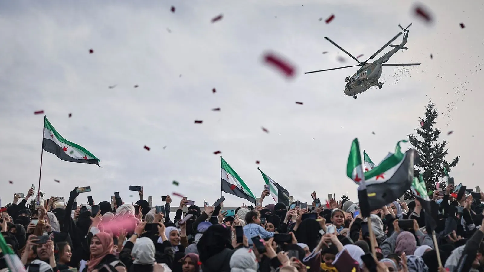 مروحية تحلق فوق حشود من المحتفلين في سوريا، حيث يرفعون الأعلام ويحتفلون بتشكيل الحكومة الانتقالية الجديدة.