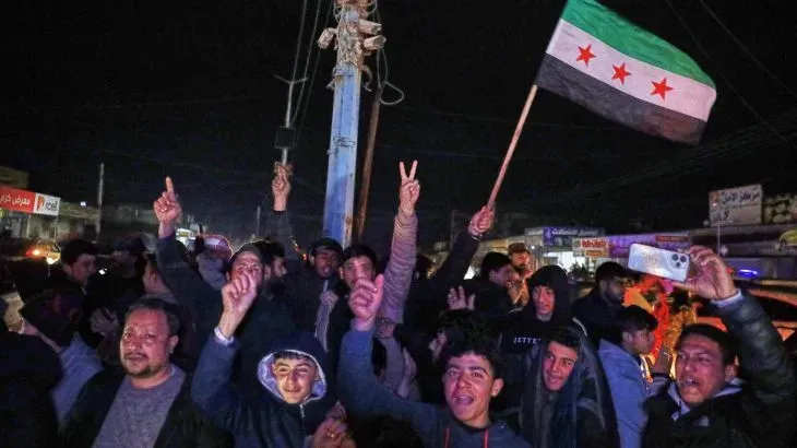 احتفال حاشد في الشارع ليلاً، حيث يرفع المتظاهرون علم سوريا ويعبرون عن فرحتهم بوقف إطلاق النار، مع إشارات النصر.