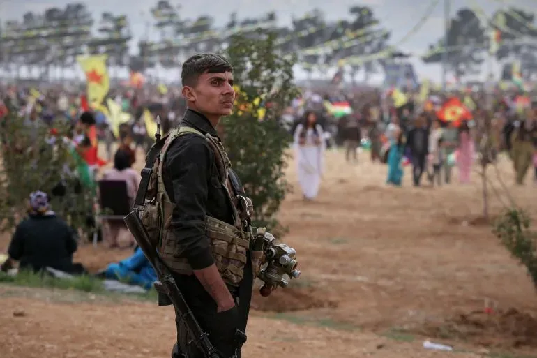 جندي من قوات سوريا الديمقراطية يرتدي زيًا عسكريًا، يقف في مواجهة تجمع حاشد في محافظة حلب، وسط توترات متزايدة مع القوات الحكومية.