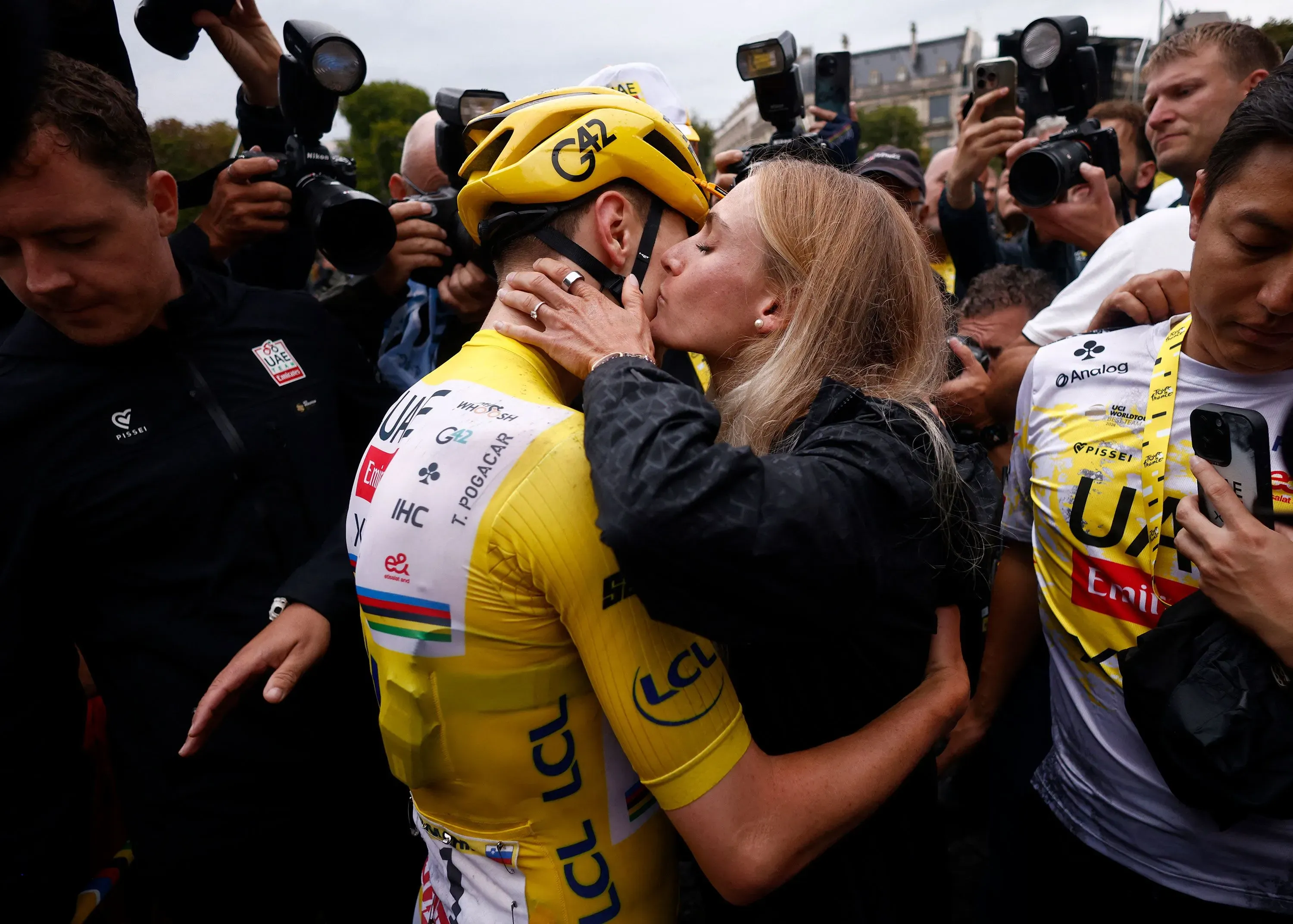 احتفال تاديج بوغاتشار بفوزه بلقب طواف فرنسا للدراجات، مرتديًا القميص الأصفر، يتلقى قبلة من شريكته وسط حشد من المعجبين.
