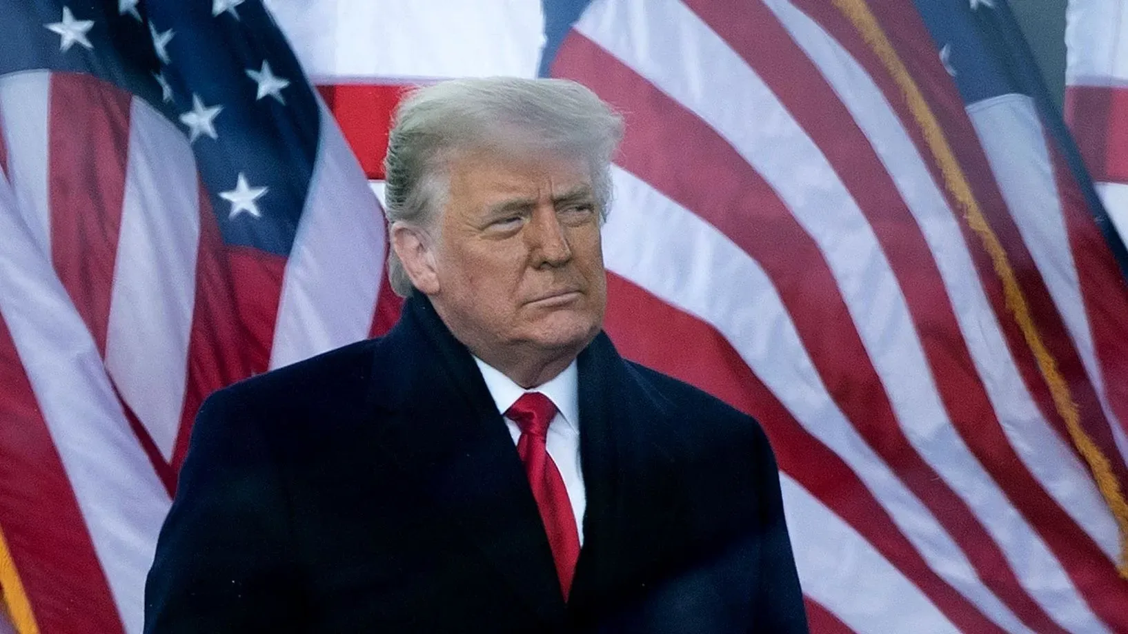 ترامب يقف أمام علمين أمريكيين، معبراً عن التوتر السياسي بعد تقرير جاك سميث حول محاولاته لإلغاء انتخابات 2020.