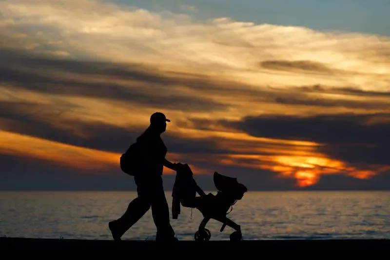 أب يتجول مع عربة أطفال على شاطئ عند غروب الشمس، مما يعكس القلق المتزايد بشأن ارتفاع أسعار مستلزمات الأطفال بسبب التعريفات الجمركية.