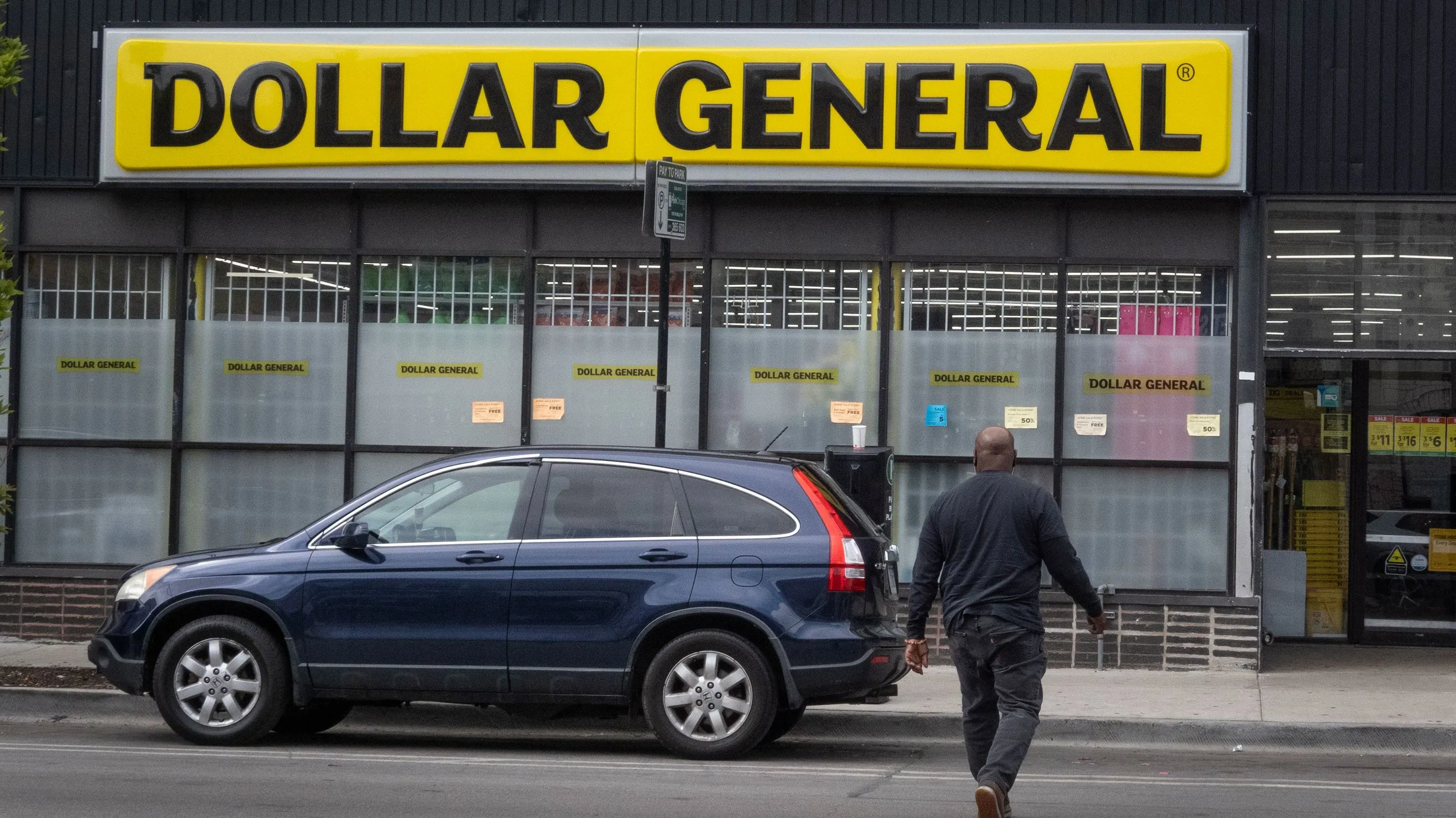 واجهة متجر "دولار جنرال" مع شعارها البارز، يظهر رجل يسير نحو المدخل، مما يعكس زيادة الإقبال بسبب الظروف الاقتصادية.