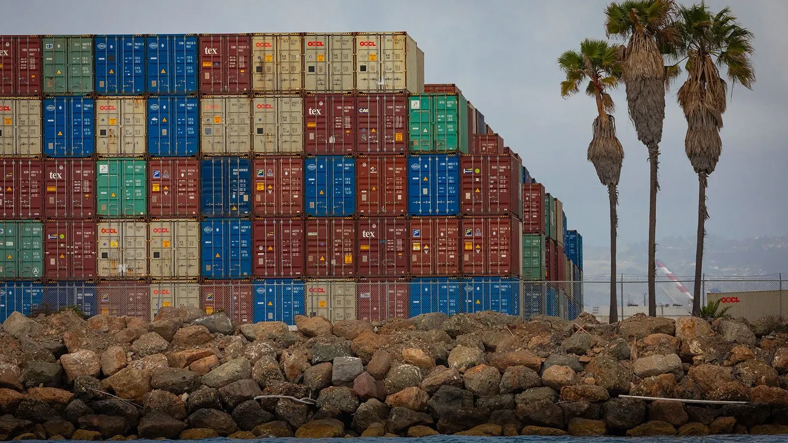 حاويات شحن ملونة متكدسة في ميناء أمريكي، مع صخور على الواجهة البحرية ونخيل في الخلفية، تعكس حركة البضائع المتأثرة بالتعريفات الجمركية.