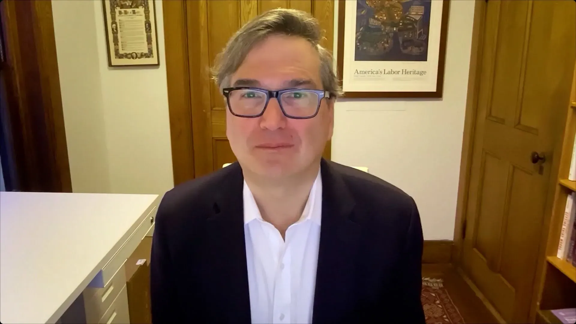 رجل يرتدي نظارات ويبتسم، يجلس في مكتب مع خلفية تحتوي على لوحات وصور تتعلق بالتراث الأمريكي.