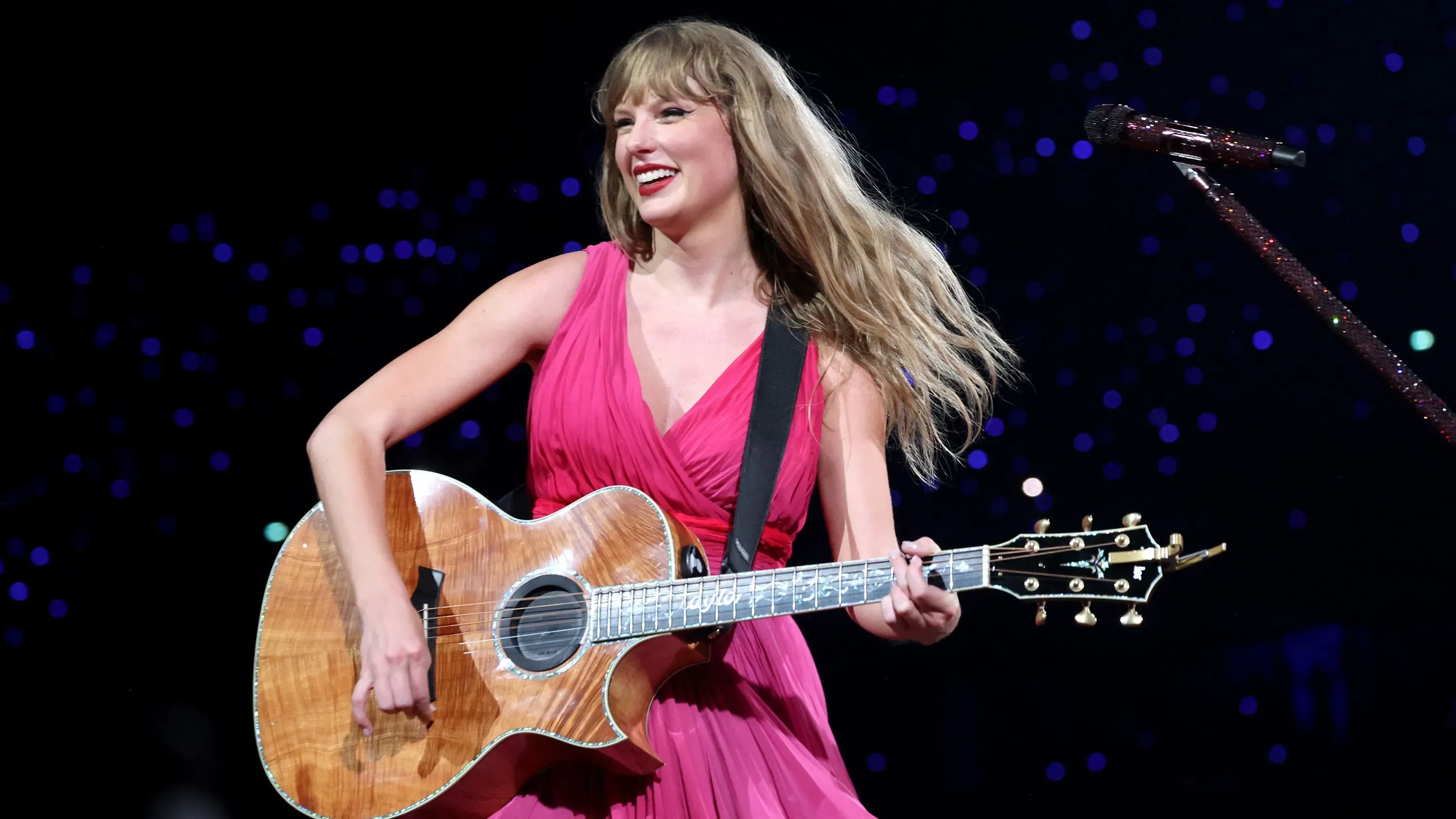 تايلور سويفت تؤدي على المسرح بفستان وردي، تحمل غيتارًا، مع أضواء خلفية ساطعة، تعبيرها يعكس فرحتها بامتلاك موسيقاها بالكامل.