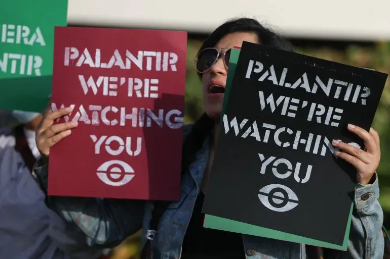 امرأة تحمل لافتتين تعبر عن احتجاجها ضد شركة Palantir، مكتوب عليهما "نحن نراقبك". تعكس الصورة القلق من تأثيرات الذكاء الاصطناعي.